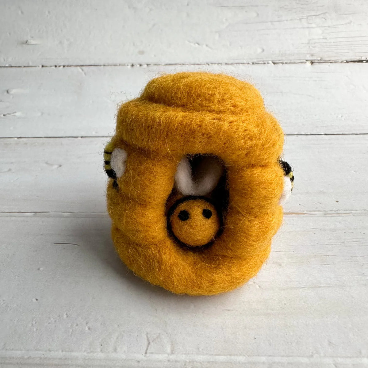 A yellow felted wool beehive toy with a small bee nestled inside, photographed on a white wooden plank surface.