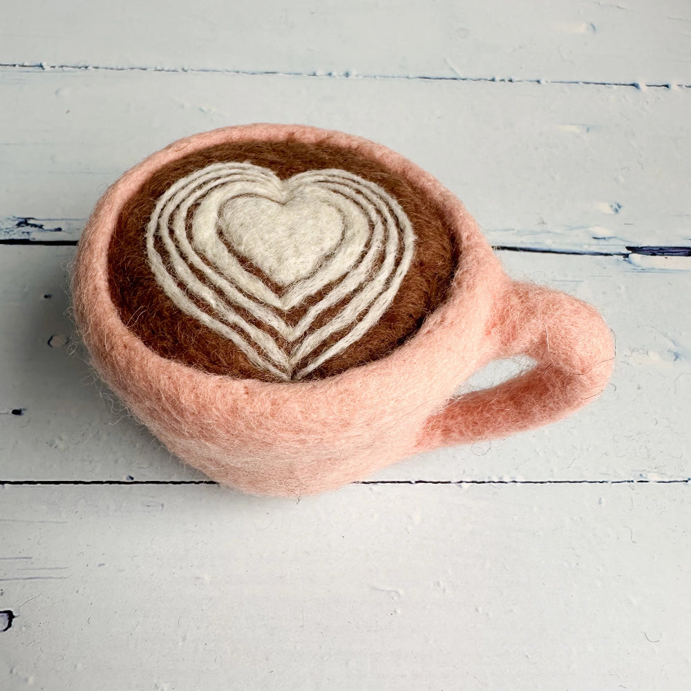 A felted wool mug shaped like a pink coffee cup with a heart-patterned latte design, resting on a white wooden surface.