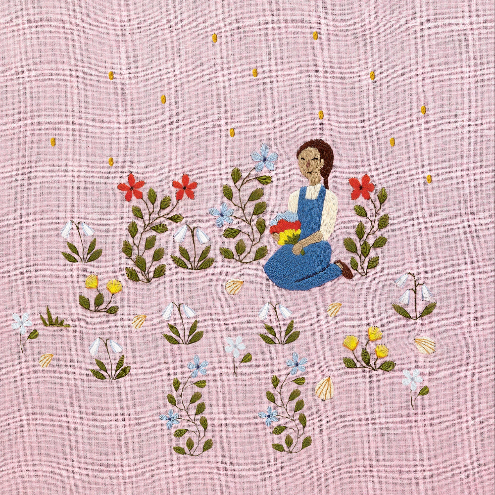 Embroidered artwork featuring a girl sitting in a field of flowers on a pink background