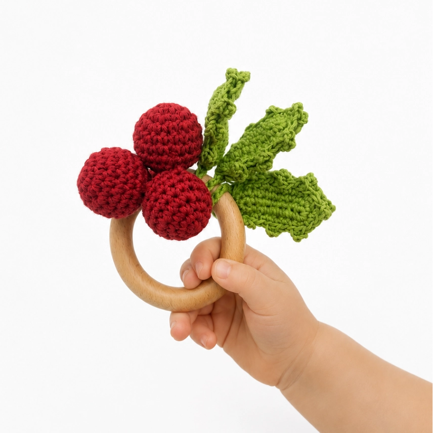Child's hand holding a wooden ring teether with crocheted holly berries and leaves on a white background