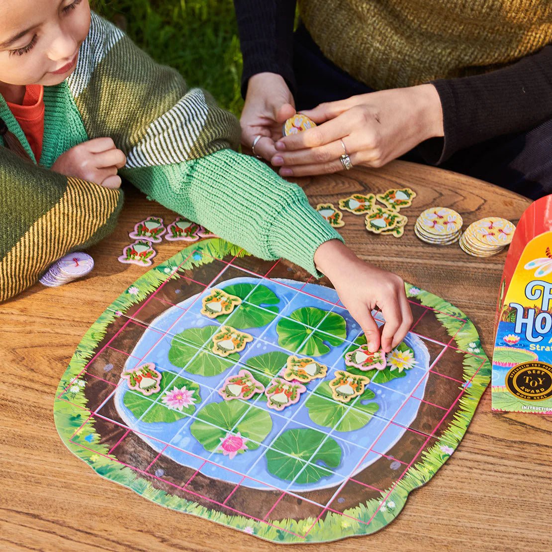 Two people playing a board game with a frog-themed board on a wooden table.