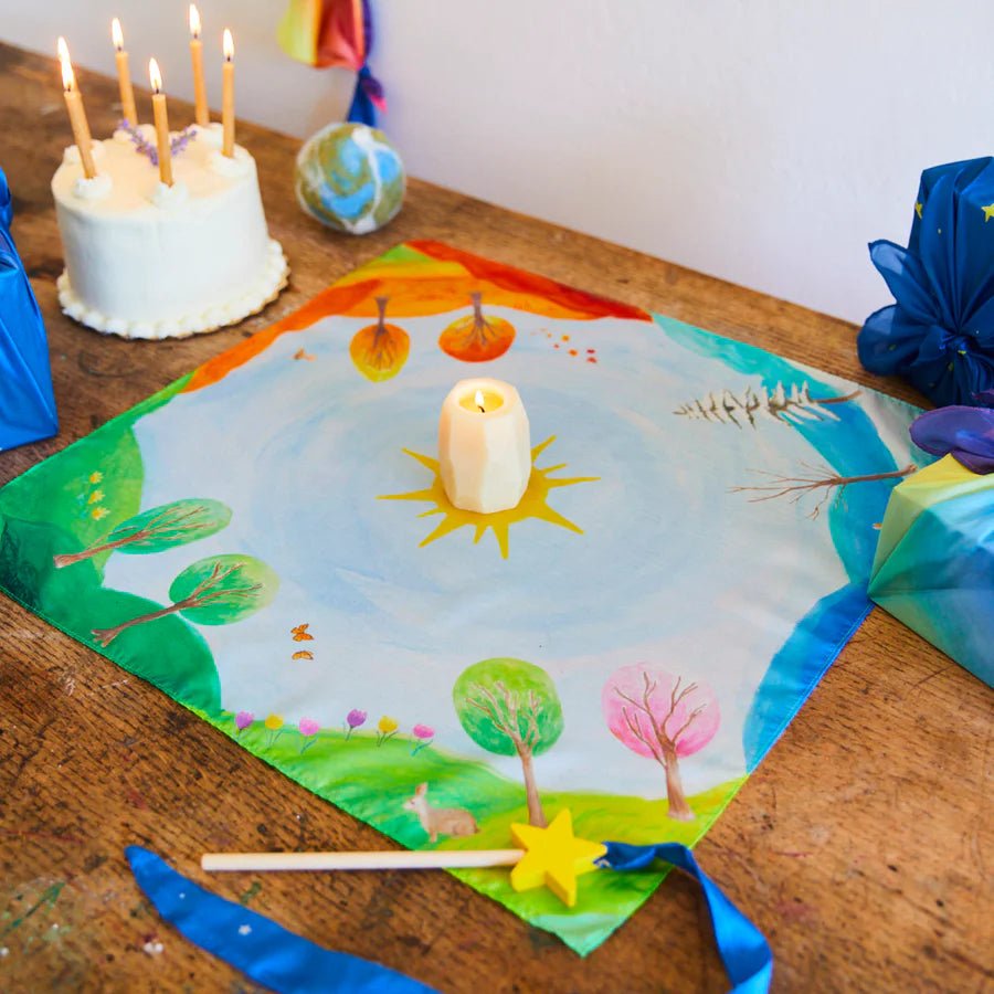 Colorful seasonal playsilk with nature design, candle, and birthday cake with candles on a wooden table.