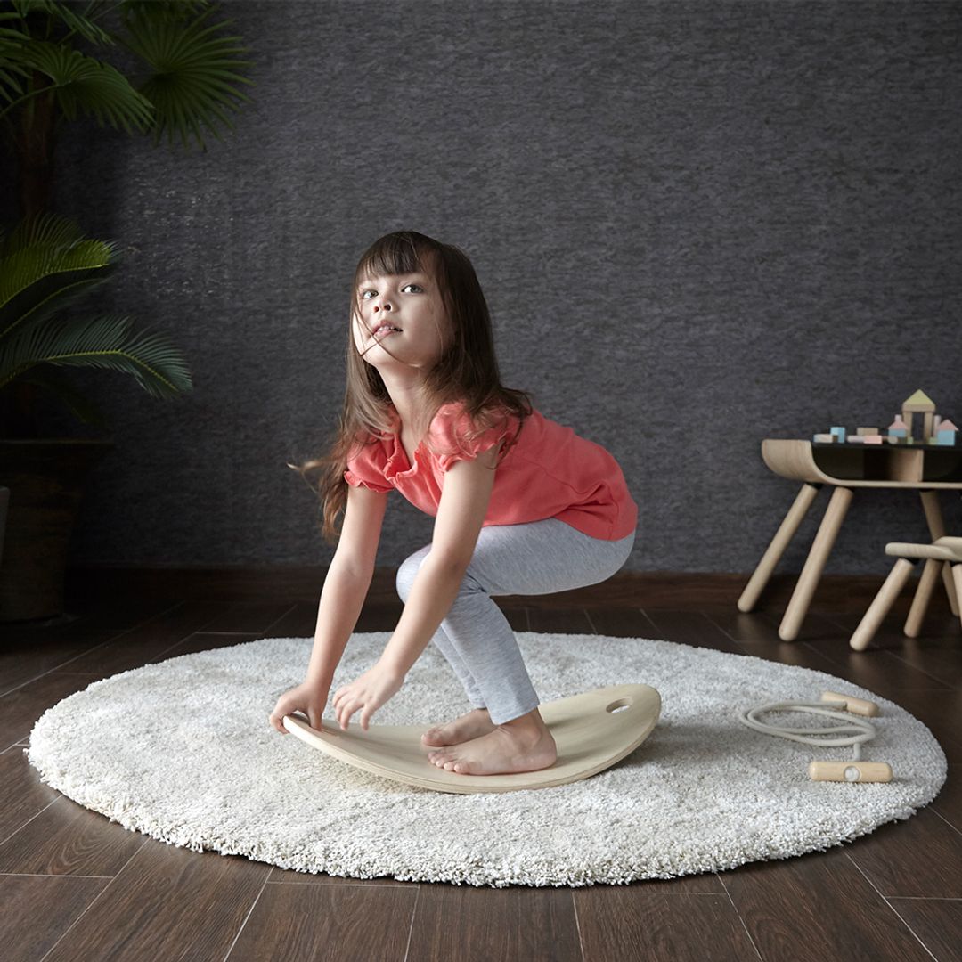 Young girl playing with a balance board on a round rug in a room with dark walls and wooden floor.