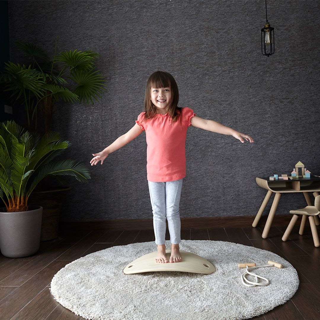 Young girl standing on a balance board in a room with a dark wall and potted plant.