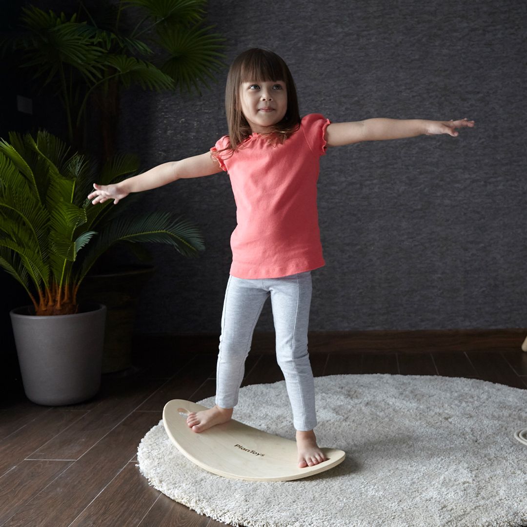 Child standing on a balance board with arms outstretched in a room with a plant and dark wall.