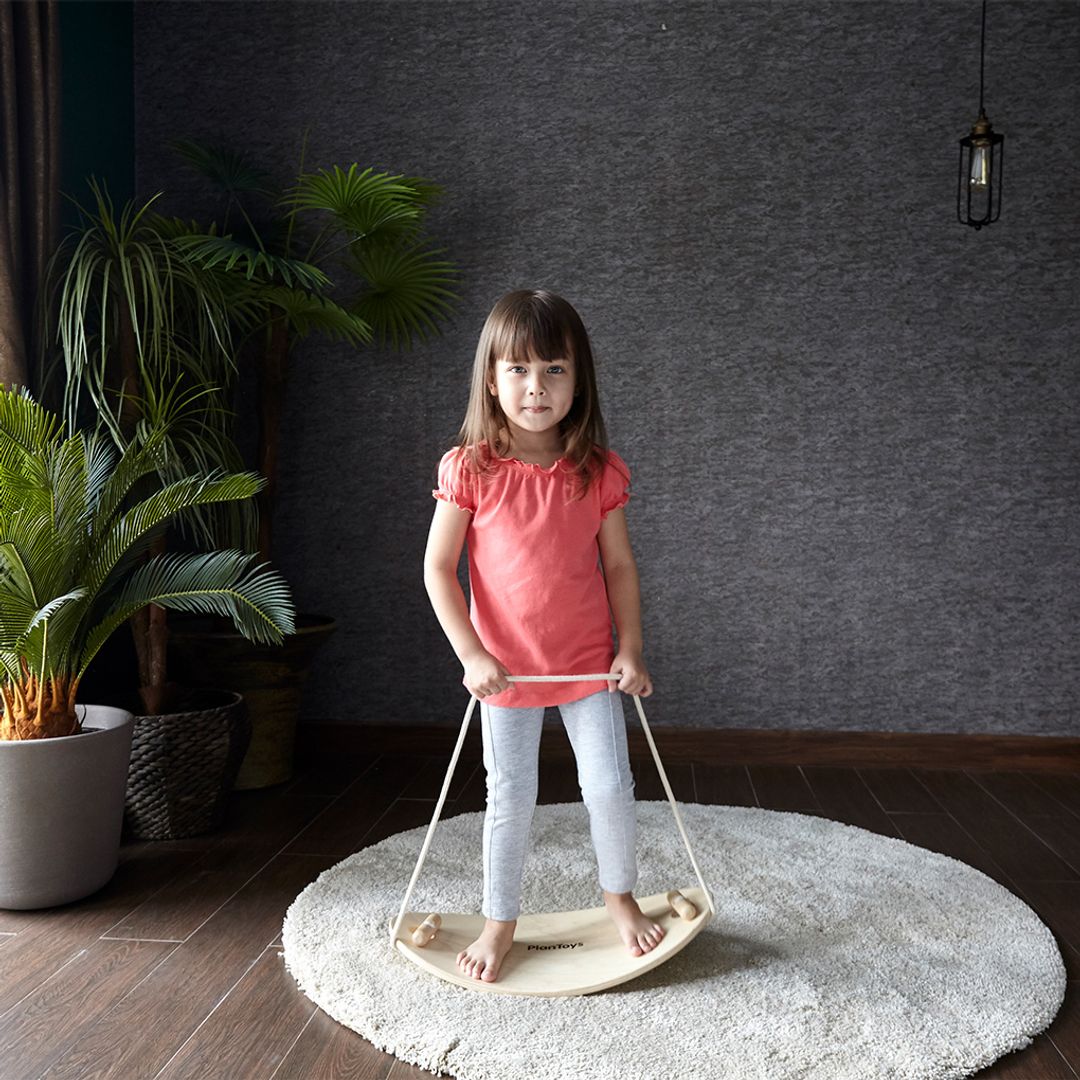 Young girl standing on a wooden balance board in a room with plants and a dark wall.