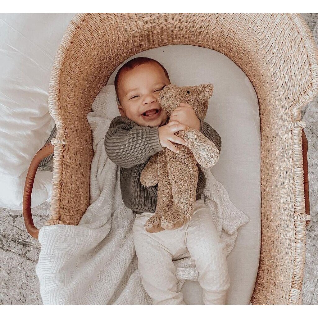 Baby holding a Senger beige teddy bear  in a wicker crib with a soft, neutral background