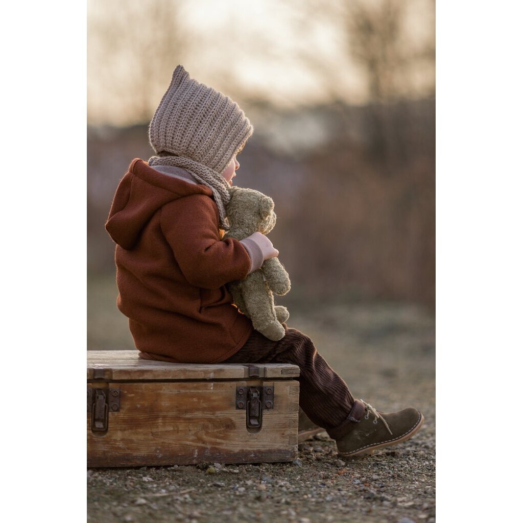 Child sitting on a suitcase holding a Senger beige teddy bear , wearing a brown coat and knit hat, with a blurred natural background.