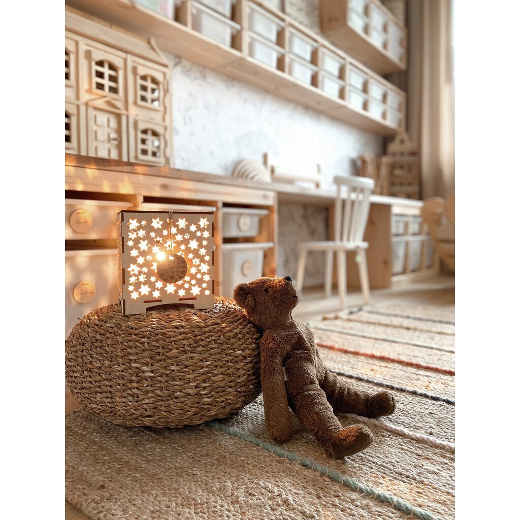 Brown Senger teddy bear sitting next to a woven ottoman with a starlit lamp in a cozy room.