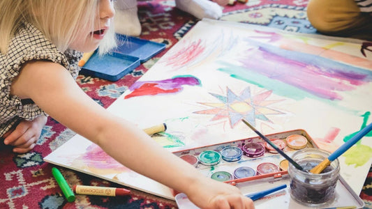 Summer Guide. A young blonde child lying on a patterned rug, reaching for art supplies next to a large sheet of colorful watercolor drawings and a paint box.