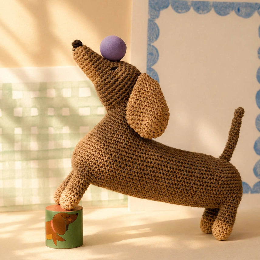 Crocheted dachshund toy balancing a purple ball on it's head with its two front feet on a platform on a light colored background