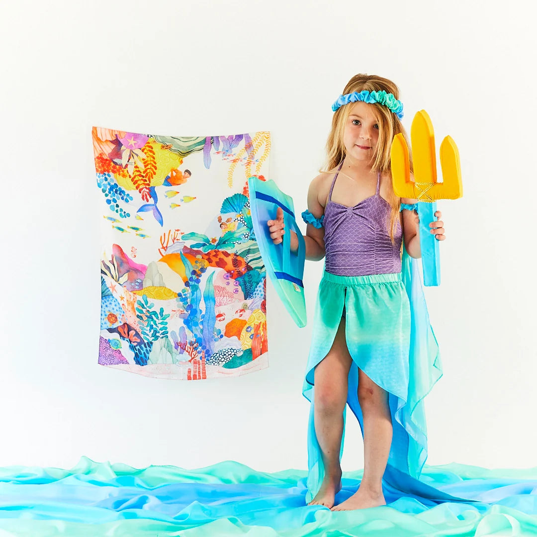 Child dressed in a mermaid costume holding a silk trident and shield, standing in front of a silk with a marine pattern