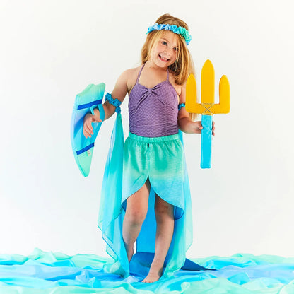 Child in a mermaid costume holding a silk shield and trident, surrounded by blue and green hombre silks against a white background 