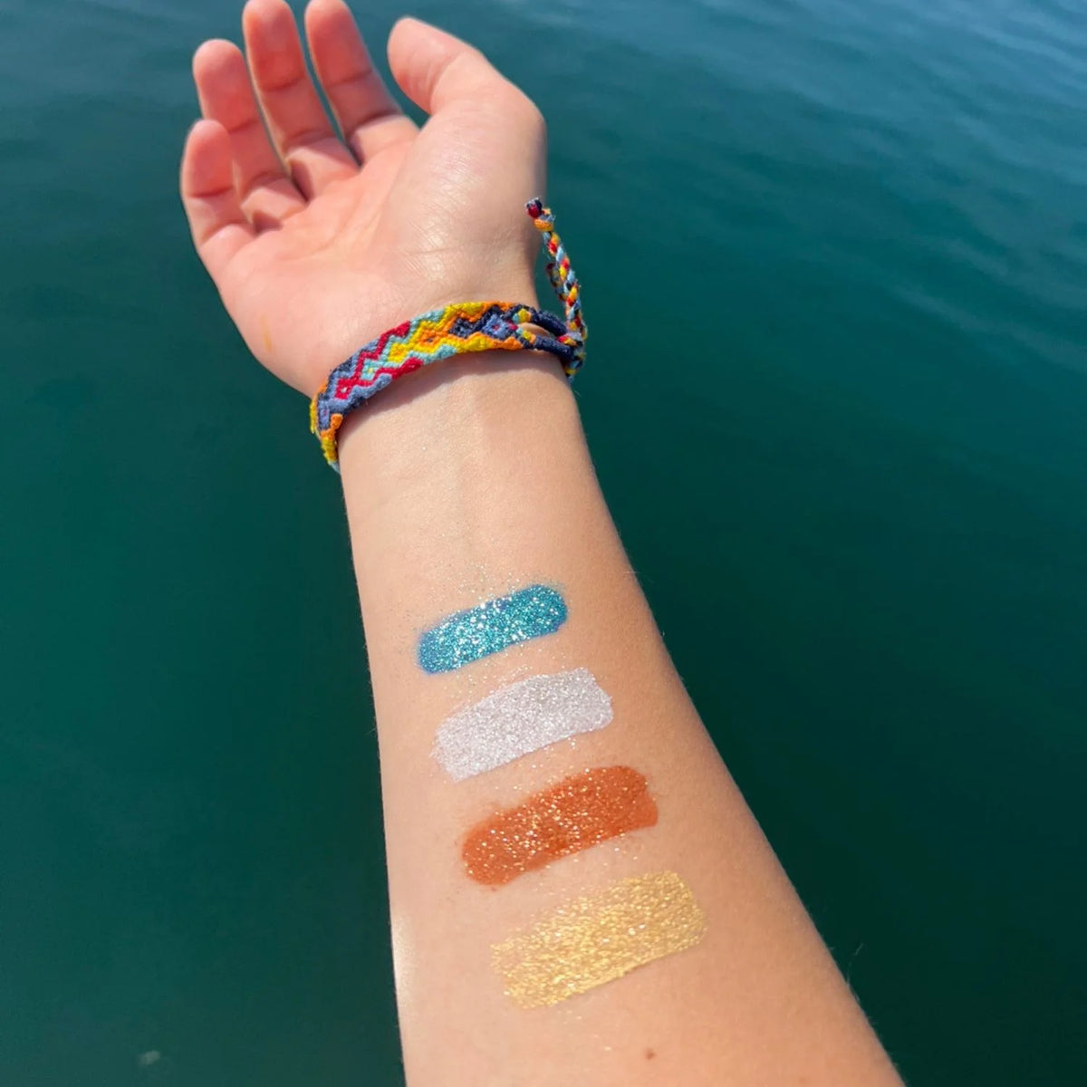 Arm with four swatches of turquoise, silver, bronze, and gold glitter with an ocean background