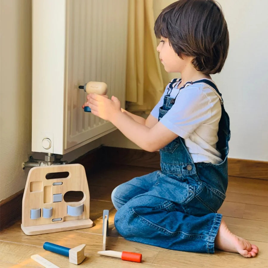 Child pretending to drill a wall radiator with a wooden drill, surrounded by other wooden tools