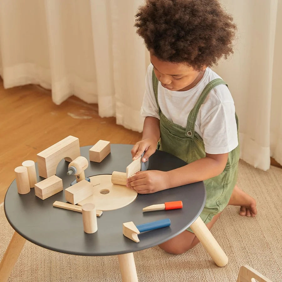 Child sitting at a table that looks like a record, with wooden tools and pieces of wood scattered on it.