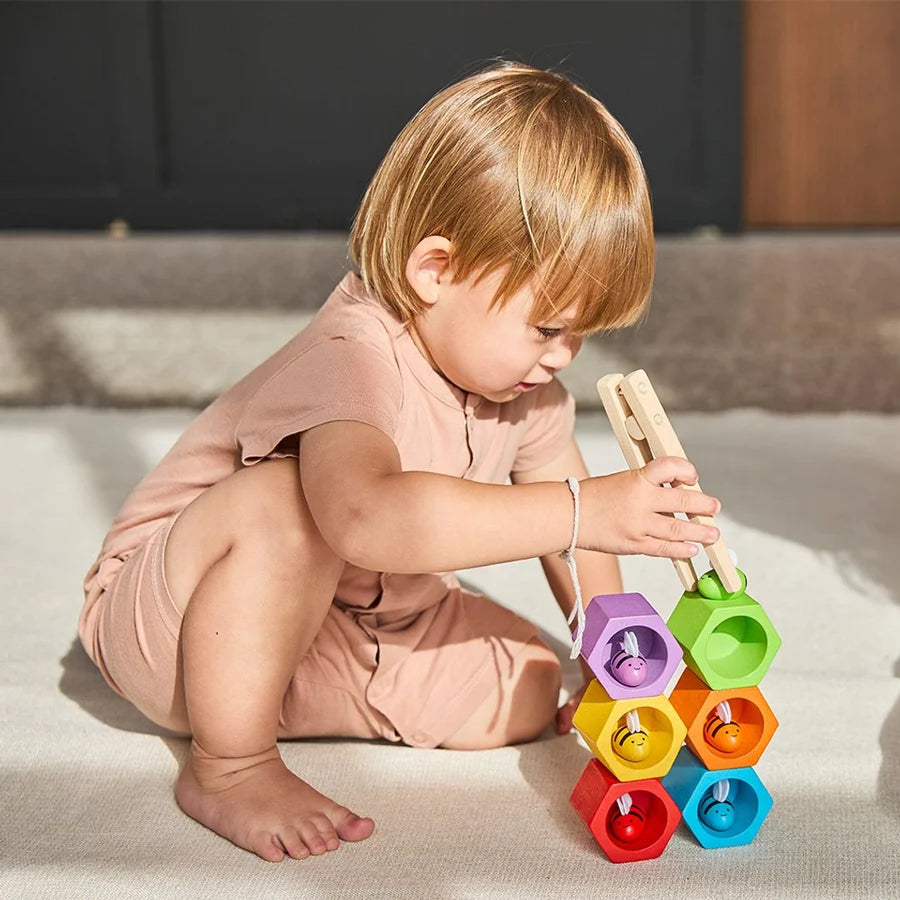 Young child using wooden tongs to place a green wooden bee on top of a stack of differently colored beehives in an outdoor setting
