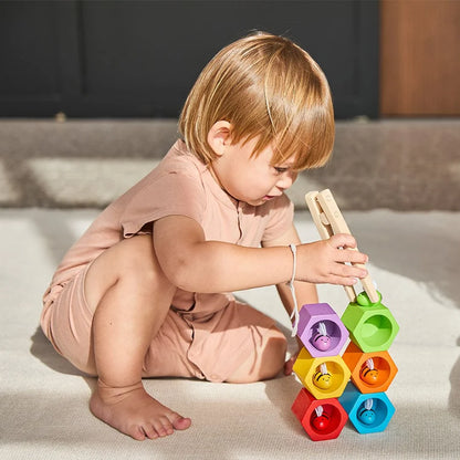 Young child using wooden tongs to place a green wooden bee on top of a stack of differently colored beehives in an outdoor setting