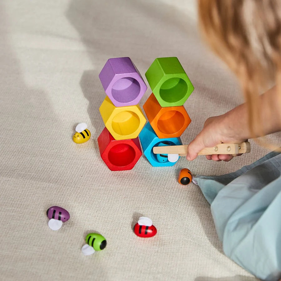 6 wooden beehives in rainbow colors stacked on top of each other while a child uses wooden tongs to place a blue bee in the blue hive