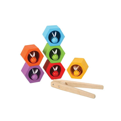 6 wooden beehives in rainbow colors with matching wooden bees stacked on top of each other with tongs on a white background