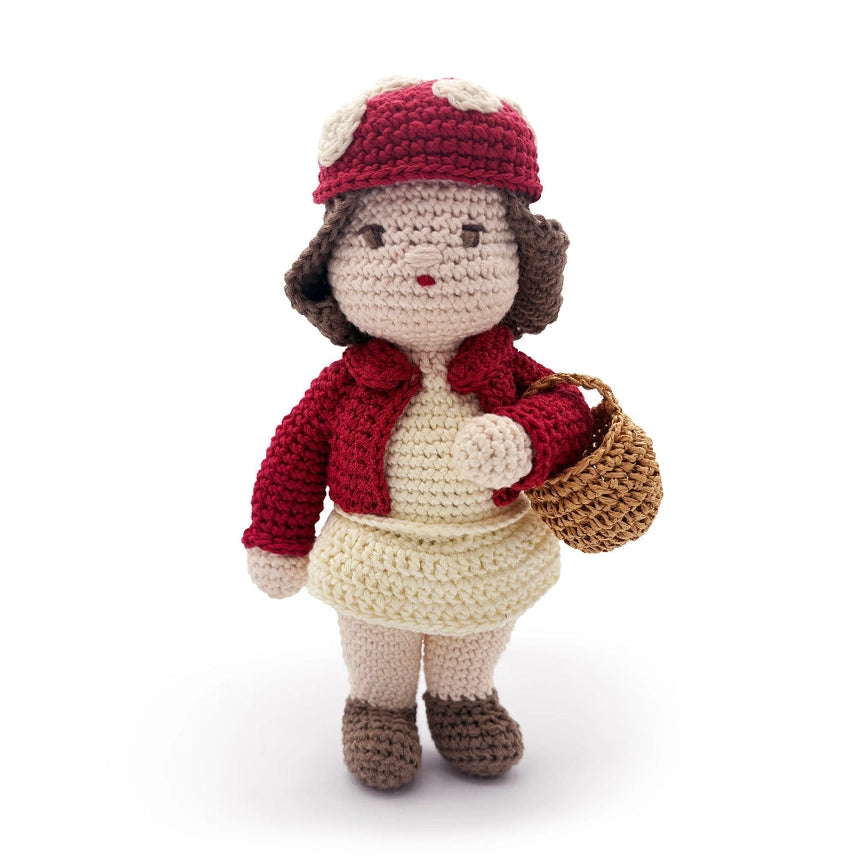 Crocheted doll wearing a beige dress, red coat, and red mushroom hat, holding a basket on a white background