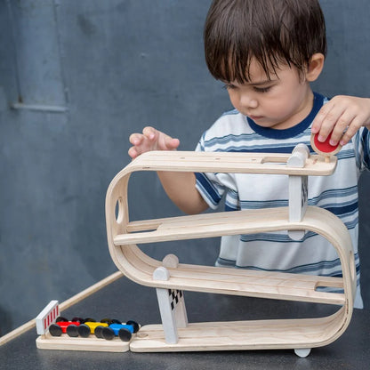 Child interacting with a wooden race track ramp and a red, yellow, and blue car at the bottom of the track.