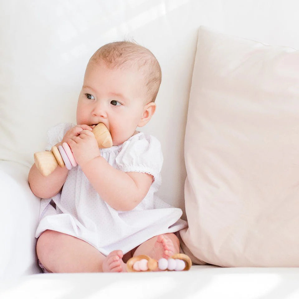 Baby in a white onesie holding a wooden baby rattle on a white couch.