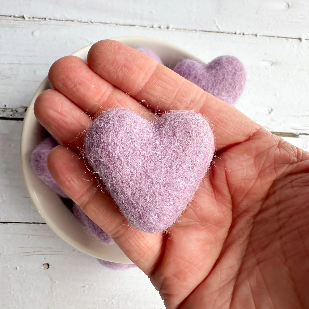 Hand holding a purple felted heart with more hearts in the background on a white wooden surface