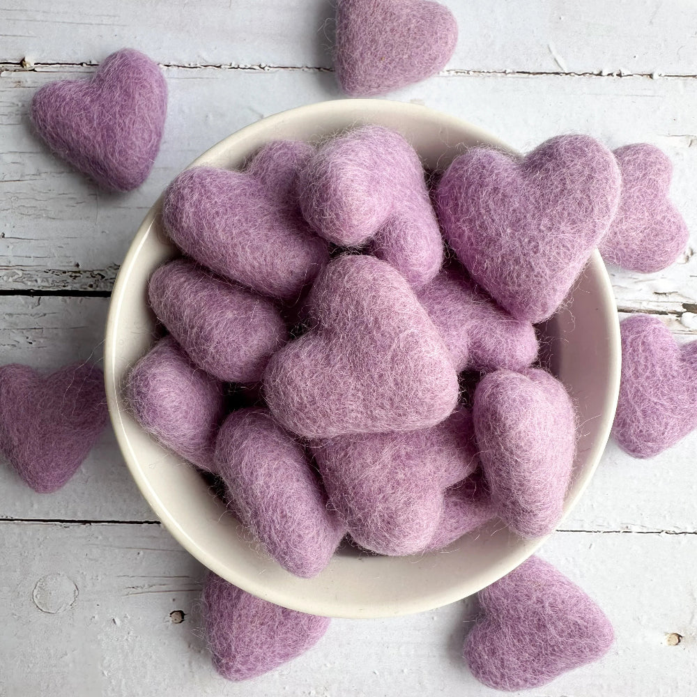 Lavender purple felted wool hearts in a white ceramic bowl and others scattered on a white rustic wooden surface