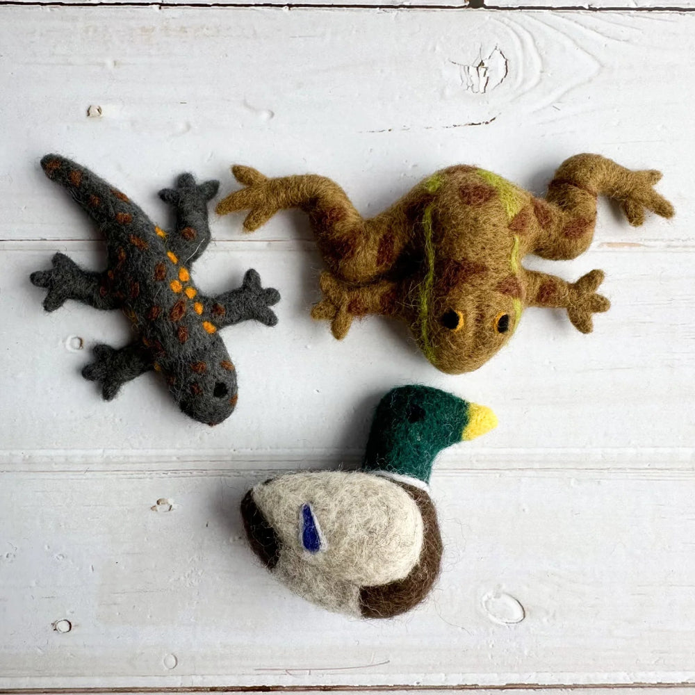 Three handcrafted felt animal toys arranged on a white wooden surface: a dark gray felted salamander with orange spots, a tan-brown felted frog with green and yellow accents, and a duck with a green head, brown body, cream body, and yellow beak.