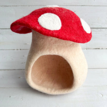 A felt mushroom-shaped doll house with a red cap, white polka dots, and a beige body, photographed against a white wood plank background.