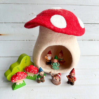 A felt mushroom-shaped gnome house with a red cap and white spots, surrounded by small painted garden gnomes and matching felt mushrooms on a white wood plank surface.