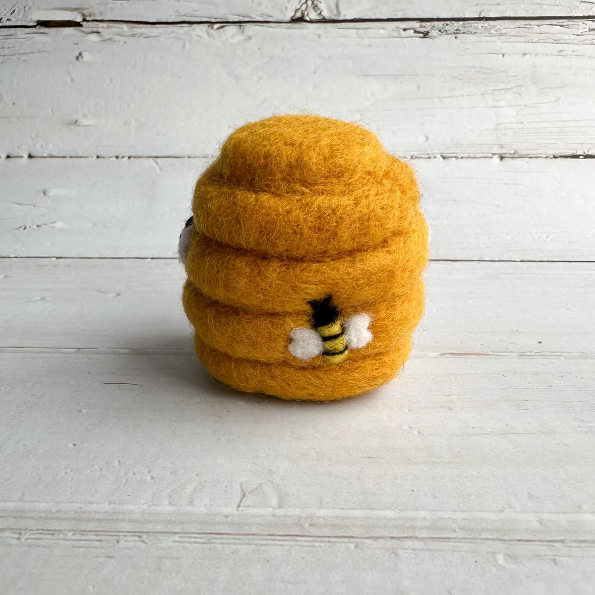 A small felted wool beehive toy in a rich golden yellow color, featuring a tiny black and yellow bee with white wings on the front, set against a rustic white wooden plank background.