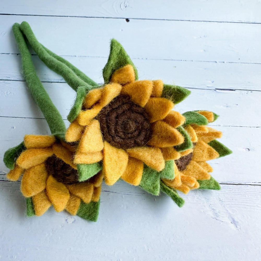 Three handmade felt sunflower stems with yellow petals, brown centers, and green leaves, arranged on a white wooden surface.