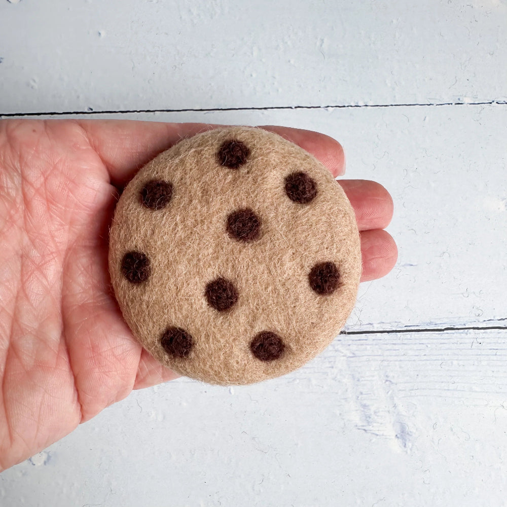 A hand holding a round felt cookie decoration with dark brown polka dots on a light beige background, set against a white painted wood surface.