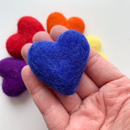 Hand holding a blue felt heart with other colored hearts in the background on a white surface