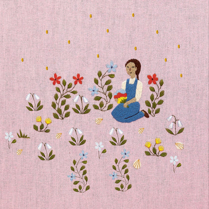 Embroidered artwork featuring a girl sitting in a field of flowers on a pink background