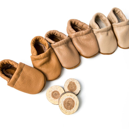 A set of soft leather baby booties in multiple tan and beige shades, arranged diagonally on a white background with three round wood slice samples below them.