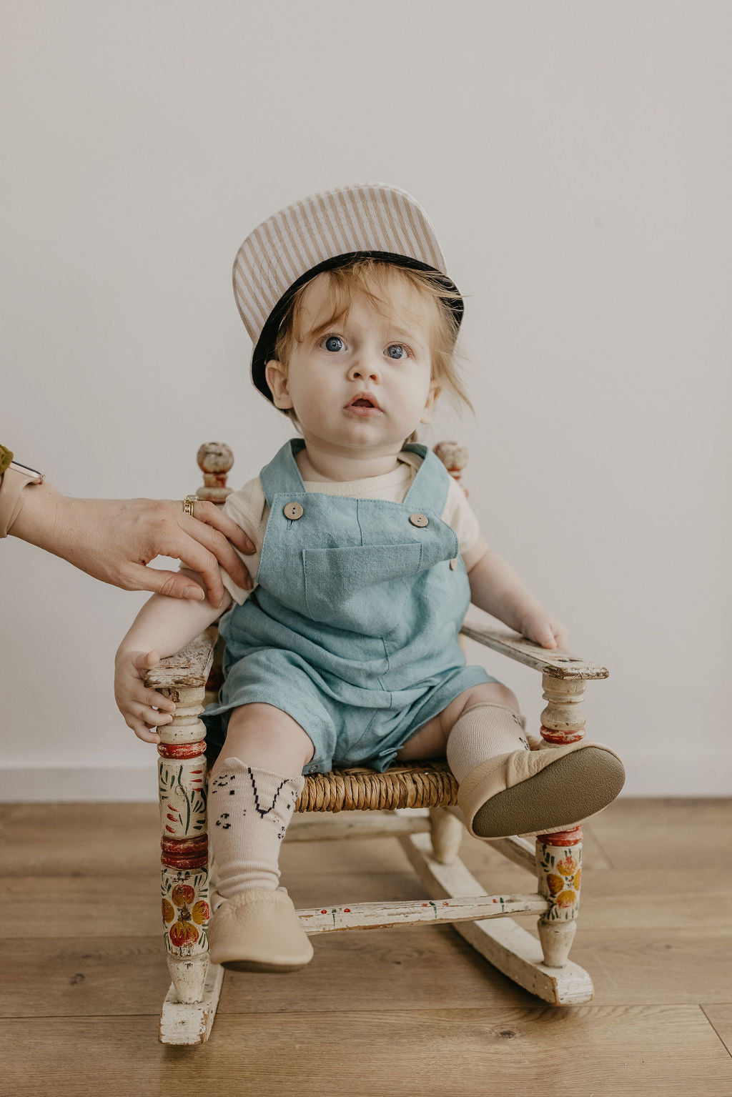 A toddler wearing a striped hat and denim overalls sits on a small decorative wooden rocking chair, with an adult hand adjusting the child's sleeve. The scene is photographed indoors against a plain light wall on a wood floor.