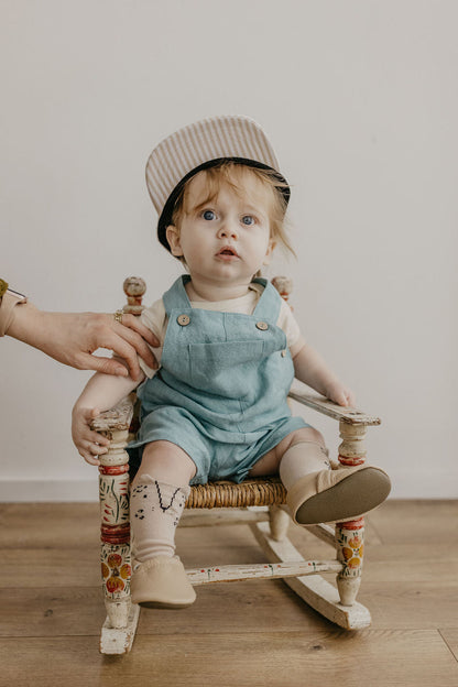 A toddler wearing a striped hat and denim overalls sits on a small decorative wooden rocking chair, with an adult hand adjusting the child's sleeve. The scene is photographed indoors against a plain light wall on a wood floor.