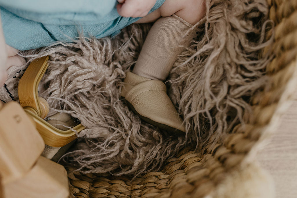 A close-up lifestyle shot of a baby's feet wearing beige knit booties resting inside a woven basket, surrounded by a fluffy gray shag rug and wooden blocks.