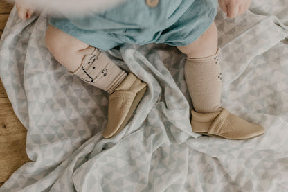 A baby wearing beige leather slip-on shoes and patterned beige socks, sitting on a light gray geometric blanket.