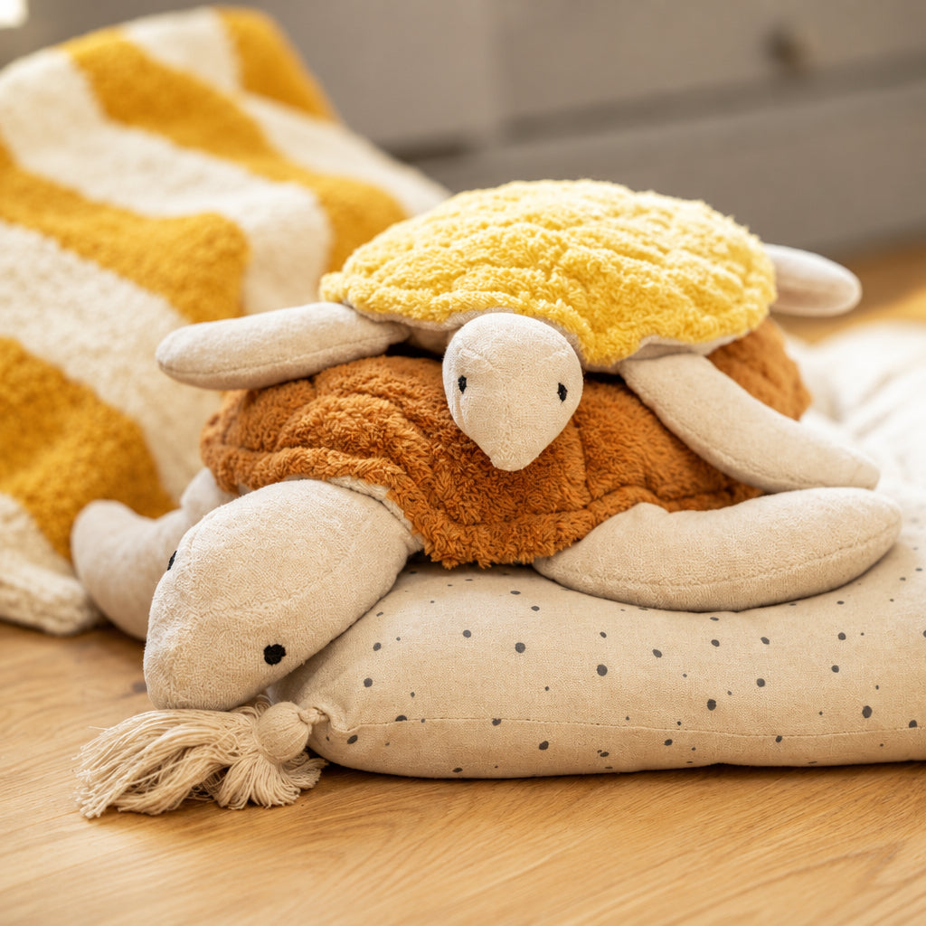 Small stuffed turtle with yellow shell on top of a larger stuffed turtle with a rust colored shell on a pillow on a wooden floor