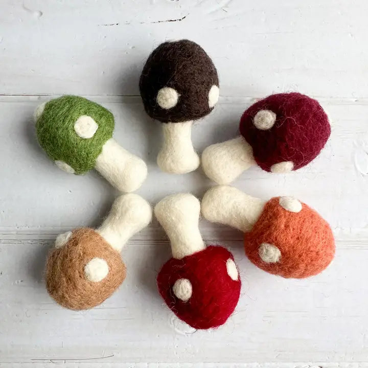 Six felt mushroom toys in assorted colors arranged in a circle on a white wooden surface. The mushrooms include green, dark brown, burgundy, orange, red, and tan, each with a white stem and white spots.