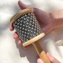 Hand holding wooden cabasa with metal beads against a neutral background