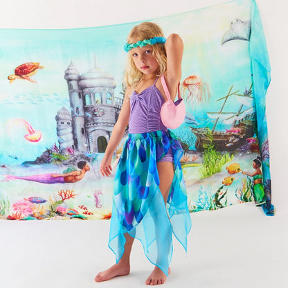 A young girl models a mermaid-themed costume set featuring a purple tank top, a blue and purple patterned high-low skirt, a matching blue headband, and a pink shoulder bag, posed in front of a large underwater castle backdrop.