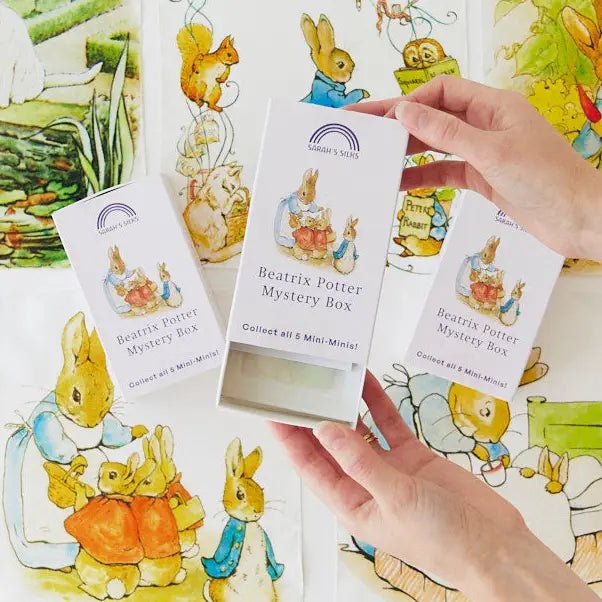 Three white Beatrix Potter Mystery Box cards are held by two hands over a background of colorful illustrated cards featuring Peter Rabbit characters.