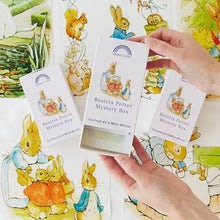 Three white Beatrix Potter Mystery Box cards are held by two hands over a background of colorful illustrated cards featuring Peter Rabbit characters.