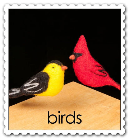 Felted yellow goldfinch and red cardinal hovering over a wooden surface with a black background