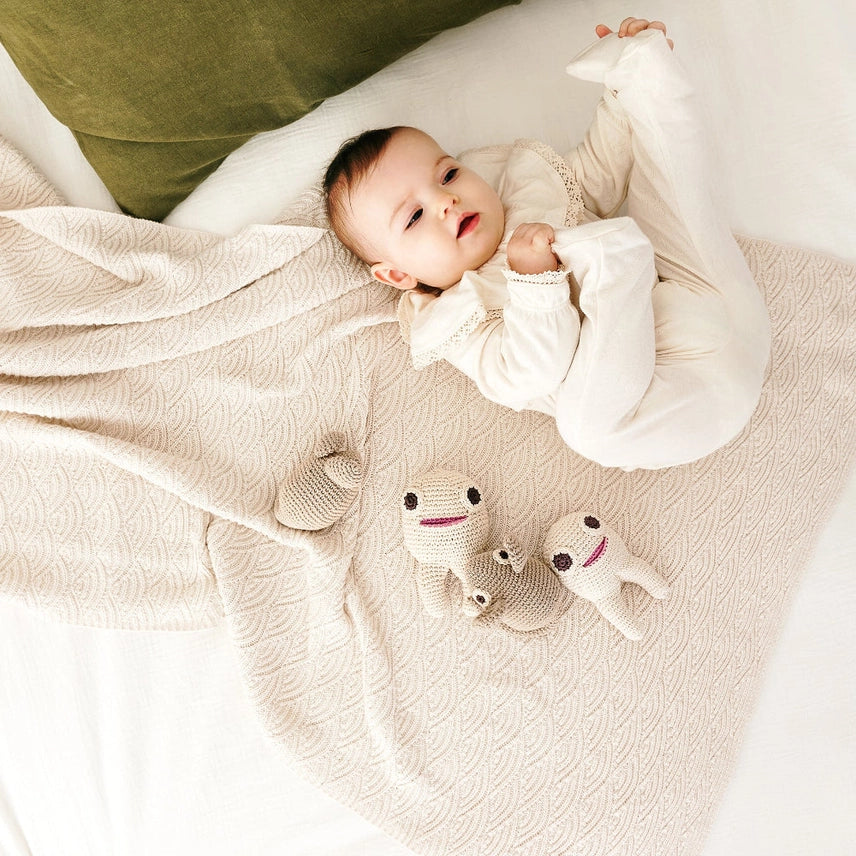 Baby laying on a bed with a green pillow and tan blanket next to two crocheted teeth-shaped and one mouse toy